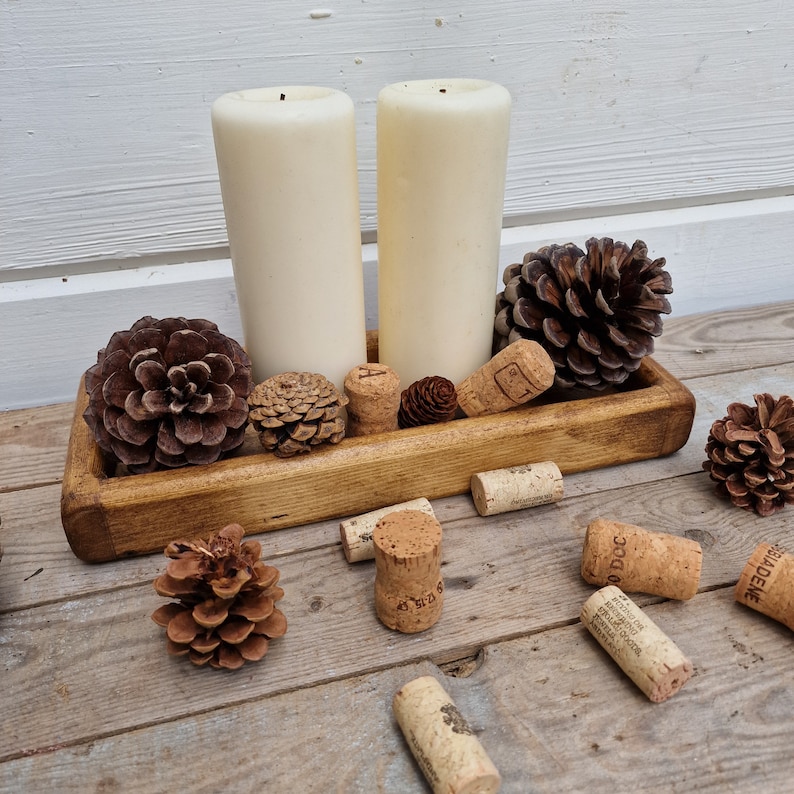 May include: A rustic wooden tray holds two ivory pillar candles, pine cones, and wine corks. The arrangement is set on a wooden surface, creating a warm, natural aesthetic. Decorative elements for home decor.