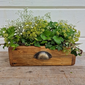 May include: A wooden planter box with a metal handle, filled with a variety of green plants and white flowers.