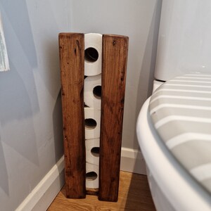 May include: A wooden toilet paper holder with four rolls of toilet paper. The holder is stained a dark brown color and has a rustic look. The holder is in a bathroom next to a white toilet.