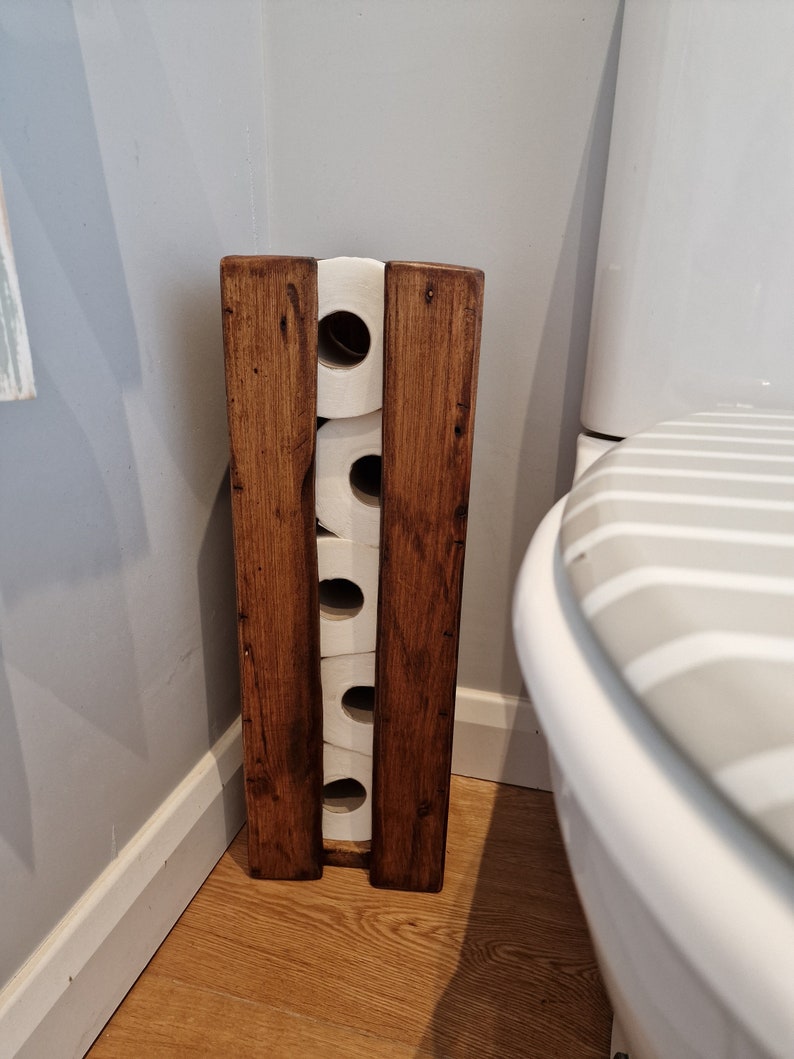 May include: A wooden toilet paper holder with four rolls of toilet paper. The holder is stained a dark brown color and has a rustic look. The holder is in a bathroom next to a white toilet.