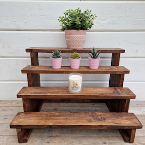 May include: A wooden tiered stand with four shelves, featuring three small potted succulents and a candle with the label "Pink Peach Blossom".