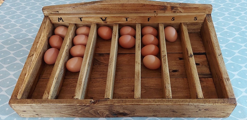 May include: A wooden egg storage box with seven compartments labeled M, T, W, T, F, S, S. The box is filled with brown eggs.