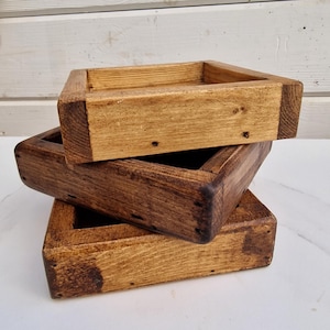 May include: Three wooden trays stacked on top of each other. The trays are made of natural wood and have a rustic, distressed look. The top tray is a lighter brown colour, while the bottom two trays are a darker brown colour.