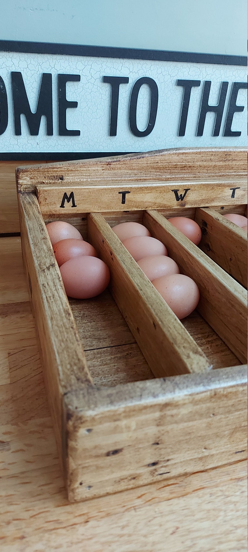 May include: A wooden egg storage box with seven compartments labeled "M", "T", "W", and "T". The box is filled with brown eggs.