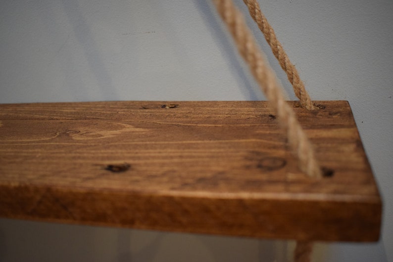K&ouml;nnte beinhalten: Ein Holzregal mit einer nat&uuml;rlichen braunen Beize, das an zwei dicken Seilen h&auml;ngt. Das Regal ist aus recyceltem Holz gefertigt und hat einen rustikalen Look.