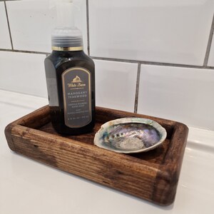 May include: A dark brown wooden tray holds a bottle of Mahogany Teakwood foaming hand soap and an iridescent abalone shell. The soap bottle has a black label with gold accents. The tray has rounded corners.