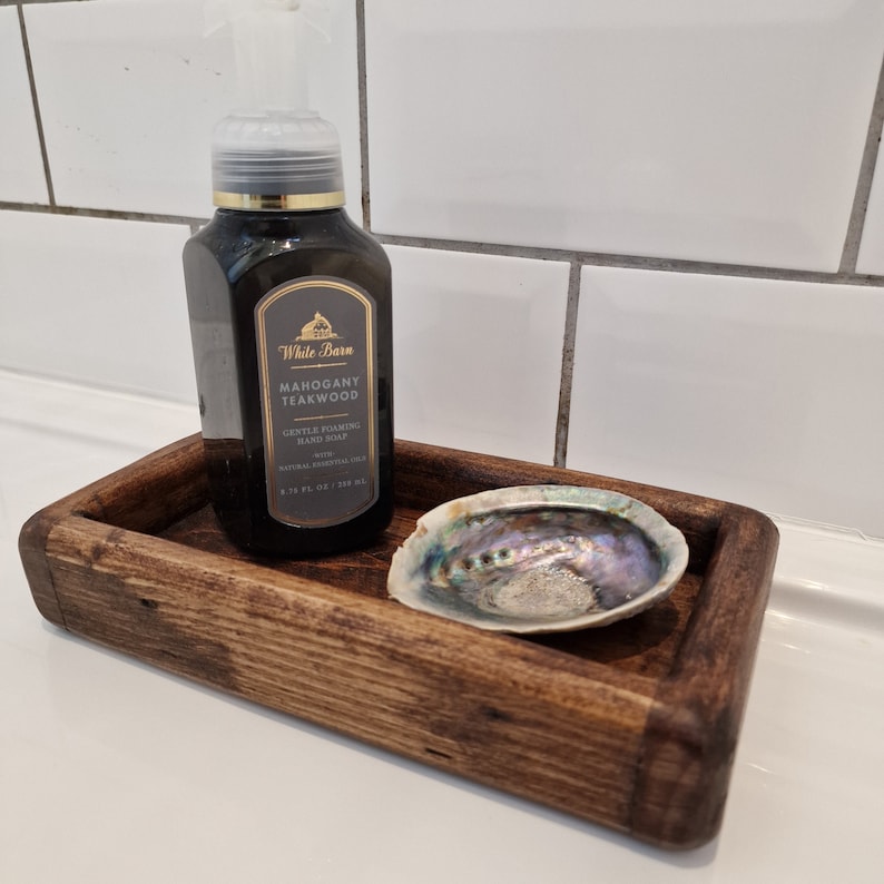 May include: A dark brown wooden tray holds a bottle of Mahogany Teakwood foaming hand soap and an iridescent abalone shell. The soap bottle has a black label with gold accents. The tray has rounded corners.