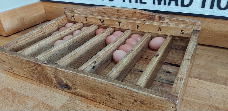 May include: Wooden egg tray with seven rows of slots for storing eggs. Each row is labeled with a day of the week, starting with "M" for Monday and ending with "S" for Sunday.