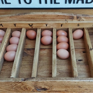 May include: A wooden egg tray with 7 slots labeled M, T, W, T, F, S, S. The tray is filled with brown eggs.