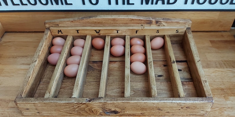 May include: A wooden egg tray with 7 slots labeled M, T, W, T, F, S, S. The tray is filled with brown eggs.