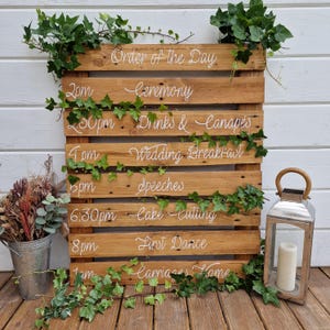 May include: A wooden pallet sign with white lettering listing the order of the day for a wedding. The sign includes the times and events for the ceremony, drinks and canapés, wedding breakfast, speeches, cake cutting, first dance, and carriages home.