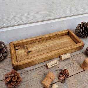 May include: A rectangular, wooden tray with a raised edge, stained in a warm brown hue. The tray is surrounded by pine cones and corks, suggesting a rustic or natural theme. The tray's dimensions are approximately 12 inches long and 6 inches wide.