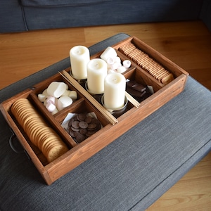 May include: A wooden tray with compartments for a s'mores party. The tray contains graham crackers, marshmallows, chocolate chips, skewers, and candles.