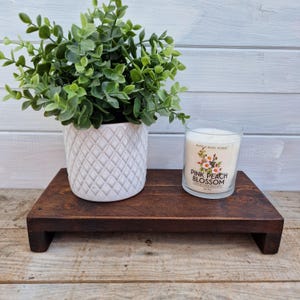 May include: A dark stained wooden riser with a white ceramic pot of greenery and a glass candle. The candle has a floral design and the text "Pink Peach Blossom". The riser is rectangular with a simple, clean design.
