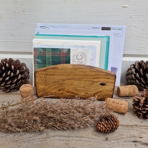 May include: A wooden letter holder with a curved top, holding a stack of envelopes and a piece of paper with a plaid pattern. The holder is sitting on a wooden surface with pine cones and cork stoppers.