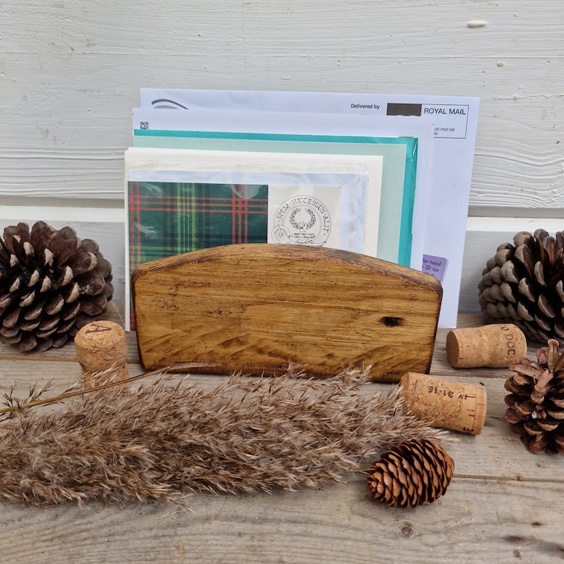 May include: A wooden letter holder with a curved top, holding a stack of envelopes and a piece of paper with a plaid pattern. The holder is sitting on a wooden surface with pine cones and cork stoppers.