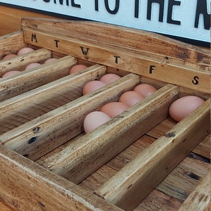 May include: A wooden egg collection box with a rustic finish. The box has seven compartments labeled with the days of the week: M, T, W, T, F, S. The box is filled with brown eggs.
