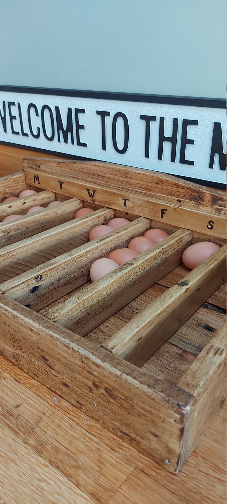 May include: A wooden egg collection box with a rustic finish. The box has seven compartments labeled with the days of the week: M, T, W, T, F, S. The box is filled with brown eggs.