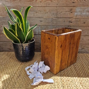May include: A rectangular wastebasket crafted from natural brown wood with a visible grain pattern, positioned next to a potted snake plant in a dark textured container and a small pile of crumpled white paper scraps on the surface.