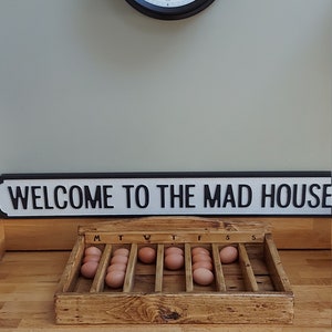 May include: A wooden egg crate with the days of the week labeled on top. The crate is filled with brown eggs. A sign above the crate reads "WELCOME TO THE MAD HOUSE".