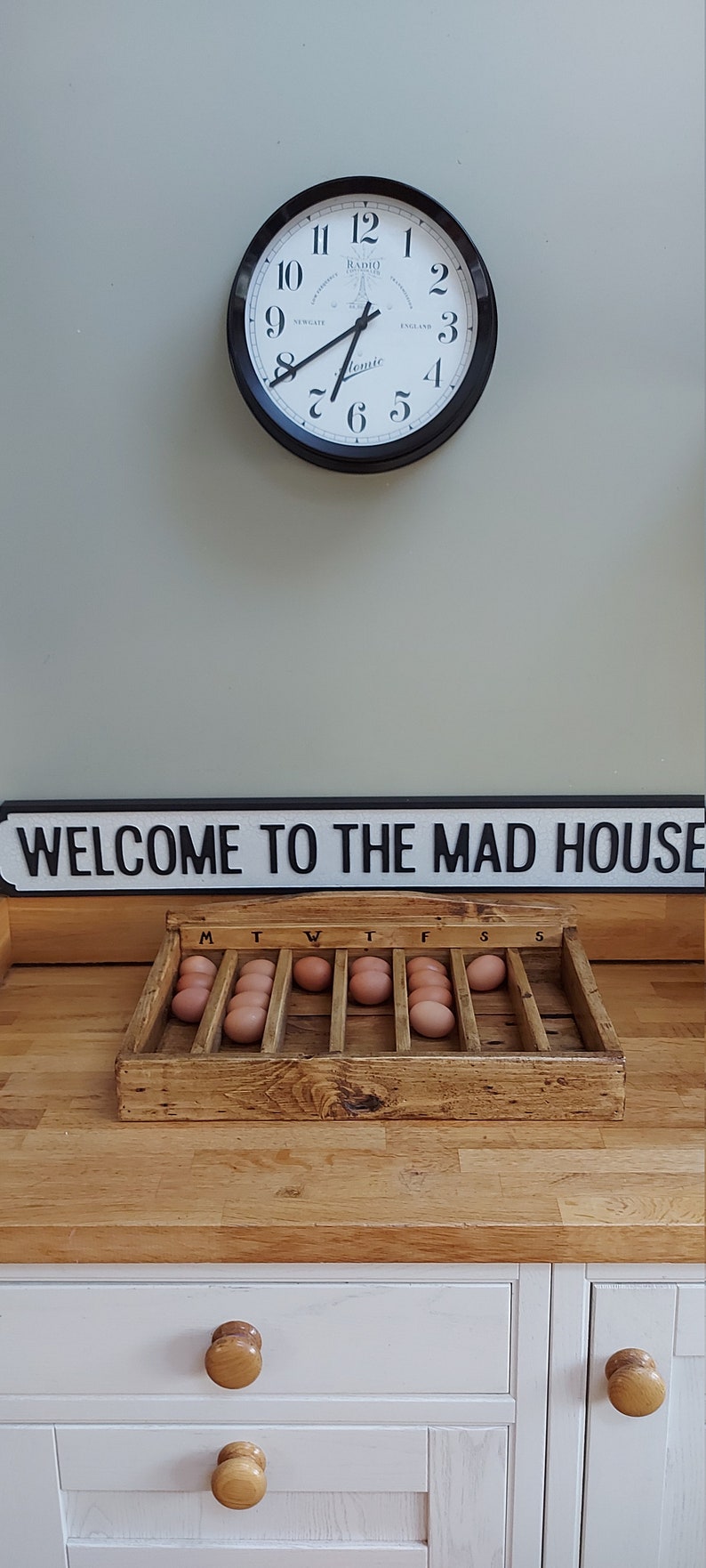 May include: A wooden egg crate with the days of the week labeled on top. The crate is filled with brown eggs. A sign above the crate reads "WELCOME TO THE MAD HOUSE".