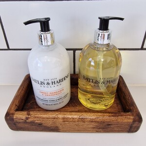 May include: Two bottles of Baylis & Harding hand wash and lotion with black pumps, on a dark wooden tray. The lotion is white with orange text, and the hand wash is yellow. The tray is rectangular and has a dark brown finish. The bottles are 16.9 fl oz each.