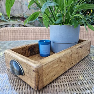 May include: A wooden tray with a metal handle, a blue candle, and a potted plant with green leaves. The tray is sitting on a wicker surface.