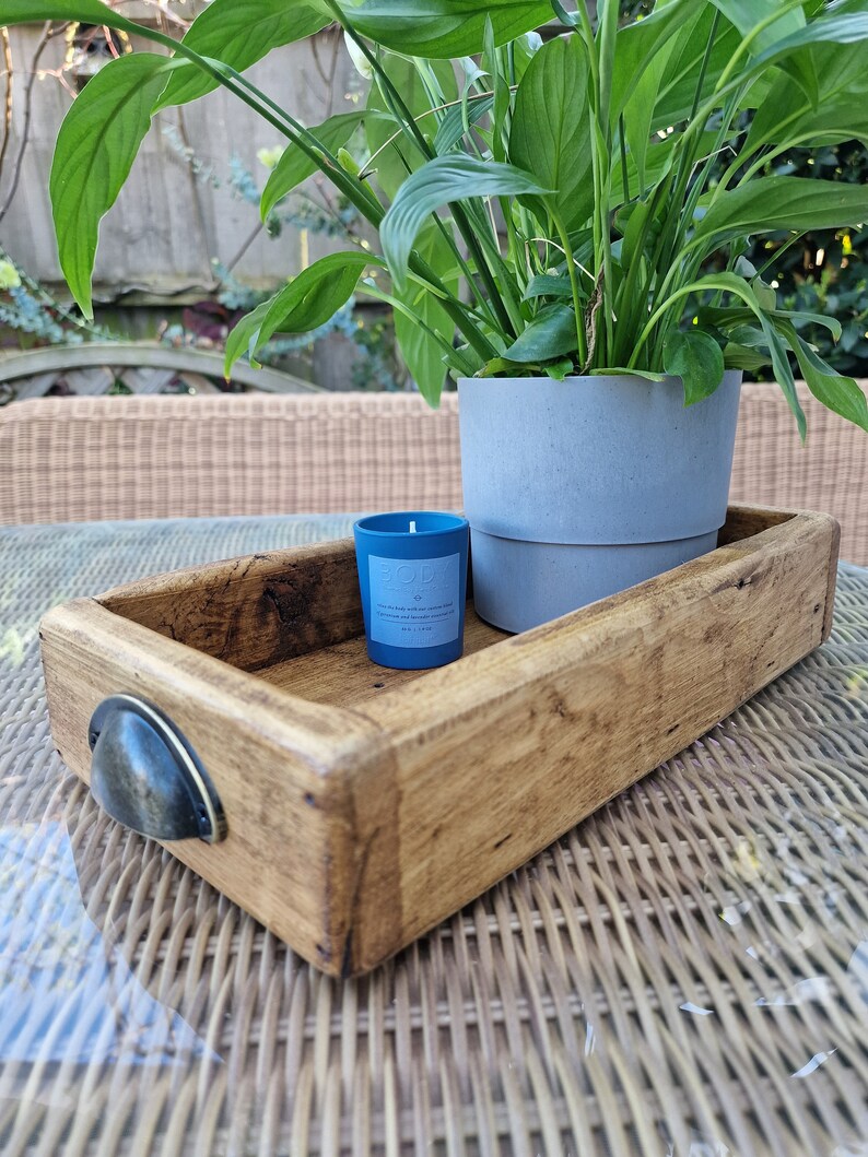 May include: A wooden tray with a metal handle, a blue candle, and a potted plant with green leaves. The tray is sitting on a wicker surface.