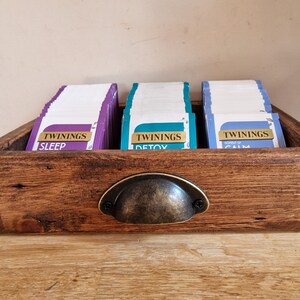 May include: A wooden tea bag organizer with a metal handle. The organizer has three compartments, each filled with tea bags. The tea bags are labeled "Twinings Sleep", "Twinings Detox", and "Twinings Power of Calm".