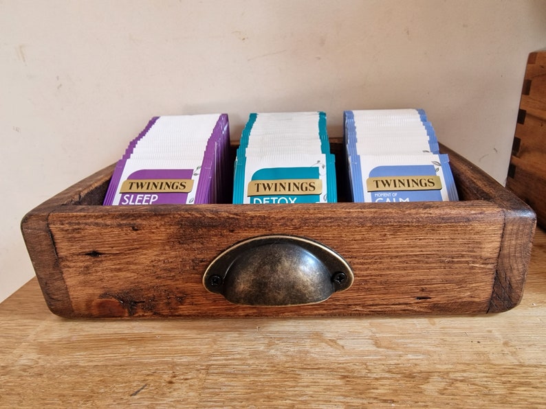 May include: A wooden tea bag organizer with a metal handle. The organizer has three compartments, each filled with tea bags. The tea bags are labeled "Twinings Sleep", "Twinings Detox", and "Twinings Power of Calm".