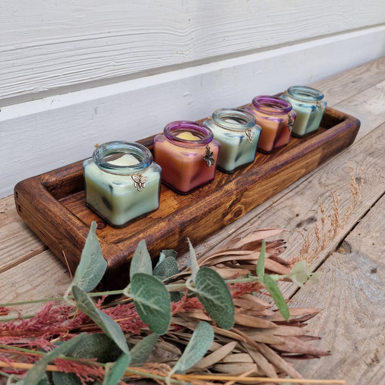 May include: A wooden tray holds five square glass jars filled with colorful candles. Each candle has a different hue, ranging from green to orange, and is adorned with a butterfly charm. The tray is stained a dark brown and sits on a wooden surface, with greenery in the foreground.