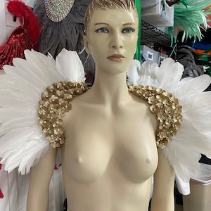 Puede incluir: Hombrera de plumas blancas con adornos florales dorados. La hombrera se exhibe en un maniquí. Los adornos dorados están hechos de pequeñas flores con pedrería en el centro. Las plumas son blancas y están dispuestas en forma de abanico.