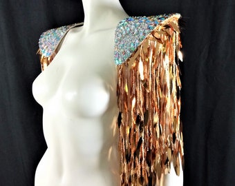 May include: A decorative shoulder piece with iridescent crystal embellishments and copper-colored fringe. The shoulder piece is displayed on a white mannequin against a black background. The fringe hangs down the arm.