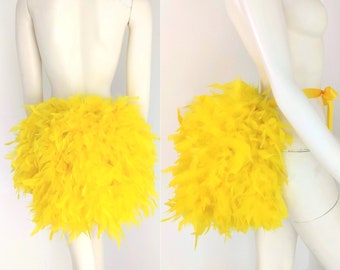 Può includere: Una gonna di piume giallo brillante, mostrata da davanti e di lato. La gonna è fatta di piume soffici e legata con un nastro giallo. La gonna è esposta su un manichino.