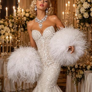 May include: A strapless white floor-length gown featuring intricate silver sequin embroidery and lace detailing. The dress is accented with large, voluminous white feather cuffs on the sleeves and is paired with a matching silver crystal statement necklace.