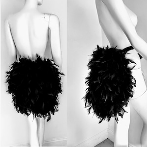 May include: A black feather skirt, displayed on a mannequin. The skirt is made of dense, fluffy black feathers, creating a voluminous, textured appearance. The skirt is designed to be worn around the waist.