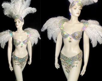 May include: A white feathered costume set featuring a bra and belt adorned with iridescent, multi-colored crystals. The ensemble includes a matching headpiece with tall white ostrich feathers and a crystal-encrusted band, worn on a mannequin.