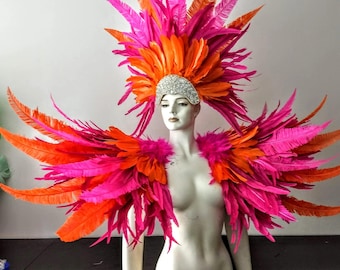 May include: A large, elaborate costume piece featuring vibrant hot pink and bright orange feathers arranged in a wide, expansive wing-like structure. A tall, matching feathered headdress sits above a silver, rhinestone-encrusted headpiece.