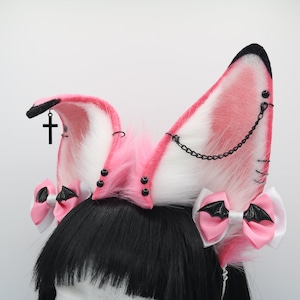 May include: Pink and white faux fur cat ears with black accents. The ears have black chains, black studs, and black bows with bat wings. A black cross charm hangs from one ear.