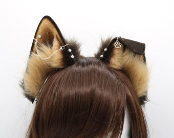 May include: A pair of brown and tan faux fur cat ears with silver chain and paw charm accents. The ears are worn on a brown wig.