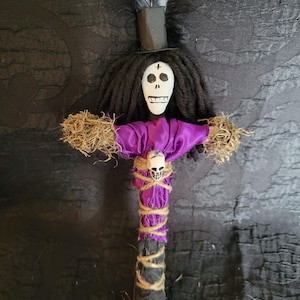 May include: A handmade voodoo doll with a painted skull face, black yarn hair, and a small top hat with a feather. The doll has a purple fabric top, a skull pendant, and is wrapped with twine.