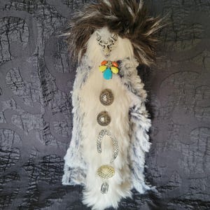 May include: A handcrafted, decorative item featuring a faux fur body in white, gray, and black. It has a detailed animal face, colorful gemstone accents, and decorative metal embellishments. A cluster of dark feathers tops the design.