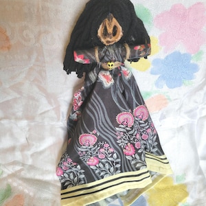 May include: Handmade doll with a black yarn wig and a carved wooden face. The doll's dress is black with pink floral patterns and a yellow border. The doll's arms are wrapped in fabric and tied with twine.