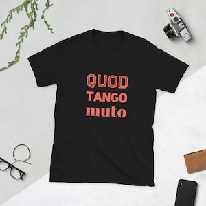 Könnte beinhalten: Schwarzes T-Shirt mit dem korallenfarbenen Schriftzug "QUOD TANGO muto". Das kurzärmelige Shirt hat einen Rundhalsausschnitt. Im Hintergrund sind eine Kamera, eine Brille und ein Telefon zu sehen.