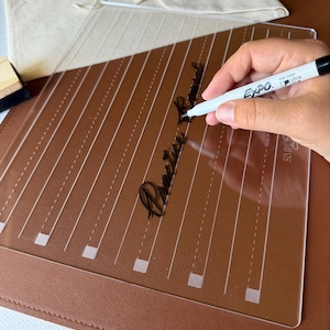 May include: A clear acrylic practice board with white printed guidelines and the text Practice Board, accompanied by a white Expo ultra fine dry erase marker, a black eraser, and a beige fabric pouch, all resting on a brown leather mat.