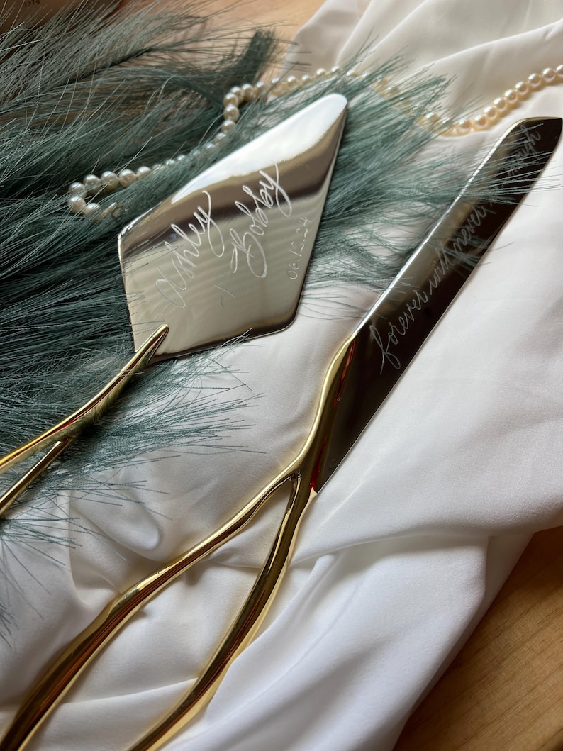 May include: A gold cake knife and server set with engraved text. The knife has the text "forever and always" and the server has the text "Ashley & Bradley 05.14.23".