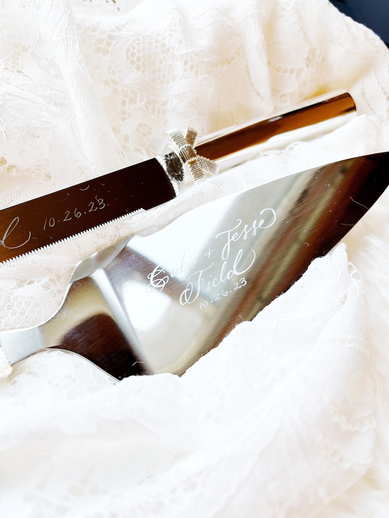 May include: Silver cake knife with a bow detail and the inscription "Gille & Jesse Field 10.26.23" on a white lace background.