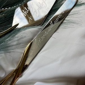 May include: A silver cake knife with a gold handle engraved with the text "Forever will never be enough". A silver cake server with a gold handle engraved with the text "Ashley + Bradley 06.12.24".