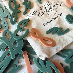 May include: A collection of matte cursive alphabet letters in muted sage green and terracotta orange, scattered around a beige drawstring fabric pouch printed with the text SIMPLY Lettered Cursive Table Set in black ink.