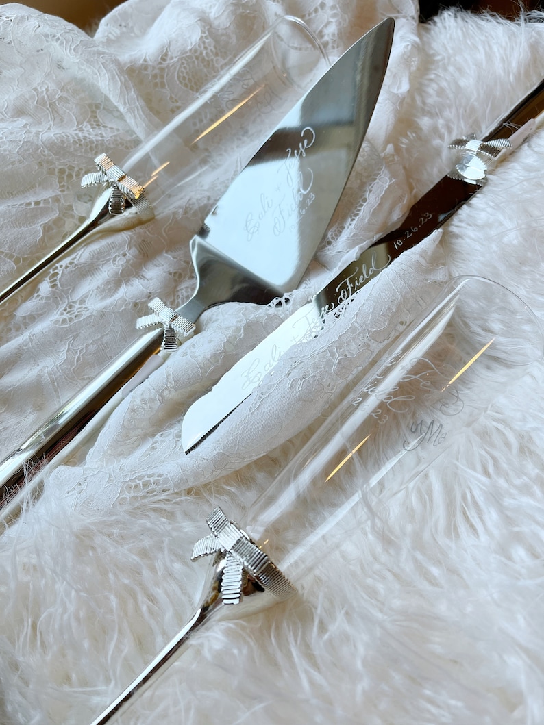May include: A silver wedding cake knife and two champagne flutes with engraved bows and the text "Emily & Jesse 10.26.23" and "Love Field Forever & Always" on the knife and "Love Field Forever & Always  & Me" on the flute.